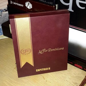 May include: A dark red wooden box with a gold label that says "La Flor Dominicana" and "CAPITULO II". The box is sitting on a wooden surface.