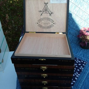 May include: A stack of wooden cigar boxes with black lids and gold accents. The top box is open and shows the brand name "Montecristo" and the text "Nicaragua Series 1935 Anniversary". The boxes are labeled "1935 Anniversary" and "10 Toro".