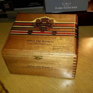 May include: A wooden box with a gold latch and a label that reads "Ashton VSG Wizard 37" and "Hand Made in the Dominican Republic". The box is made of cedar wood and has a natural finish.