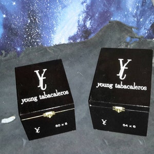 May include: Two black wooden boxes with gold hinges and the text "Young Tabacaleras" printed in white. The boxes are labeled with the dimensions "80 x 5" and "64 x 6".