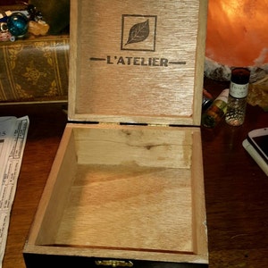 May include: A wooden box with a black lid and gold lettering. The lid is open and the inside of the box is visible. The box is labeled "L'ATELIER" and "15 LAT 52".