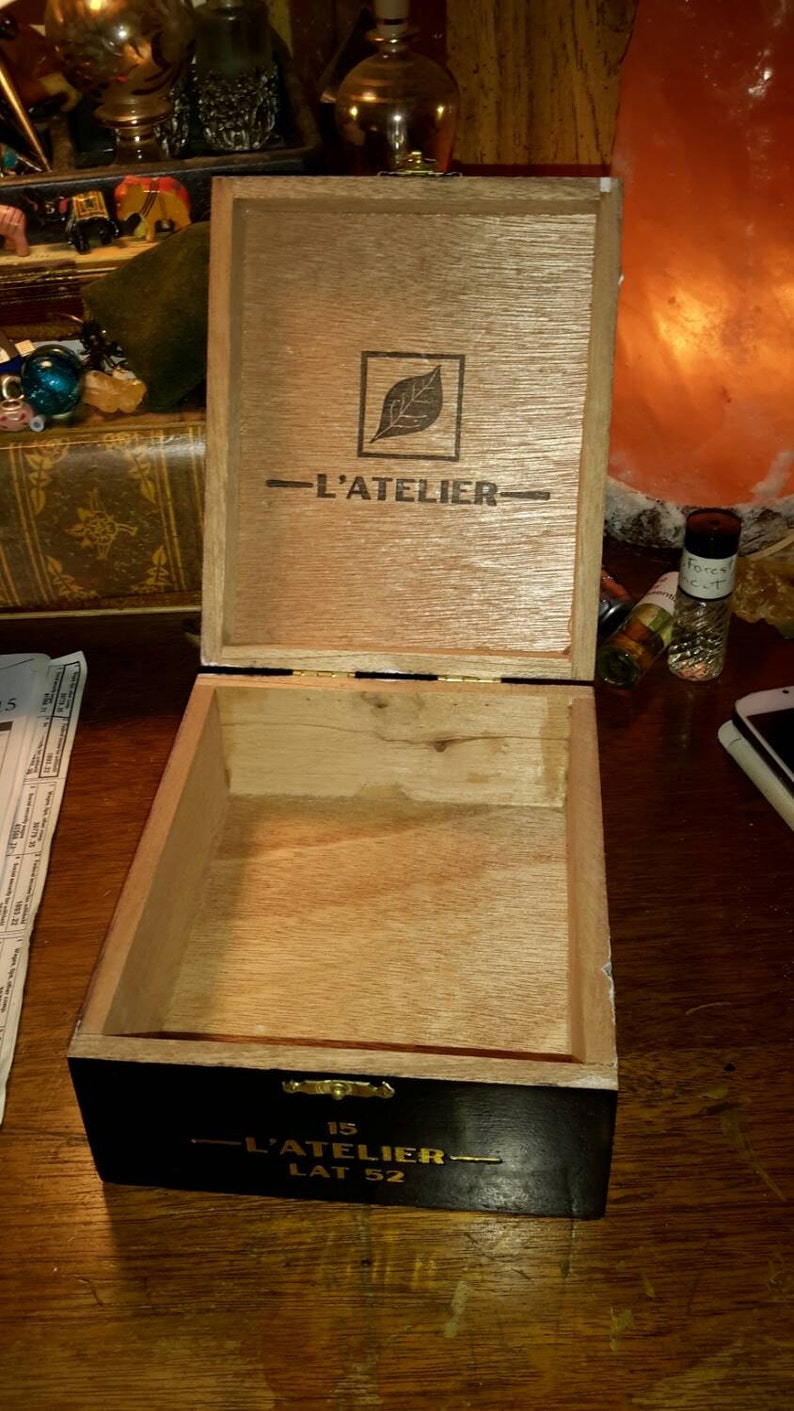 May include: A wooden box with a black lid and gold lettering. The lid is open and the inside of the box is visible. The box is labeled "L'ATELIER" and "15 LAT 52".