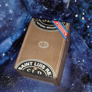 May include: A wooden box with a black label that reads "Saint Luis Rey" and "Natural Broadleaf." The box has a red, white, and blue sticker that reads "Cuban Hand-Rolled Cigars" and "Proudly Grown in the USA."