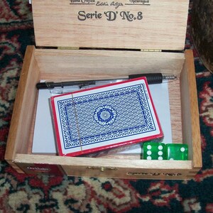 May include: An open wooden box with a blue and white playing card, two green dice, and a black pen. The box has the text "Serie 'D' No. 8" on the top.
