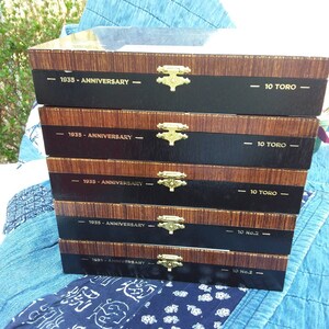 May include: Five wooden cigar boxes stacked on top of each other. The boxes are brown with black trim and gold hinges. Each box has the text "1935 - Anniversary" and "10 Toro" or "10 No.2" printed on the front.