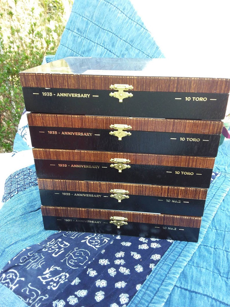 May include: Five wooden cigar boxes stacked on top of each other. The boxes are brown with black trim and gold hinges. Each box has the text "1935 - Anniversary" and "10 Toro" or "10 No.2" printed on the front.