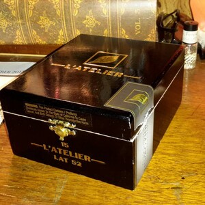 May include: A black wooden box with gold accents and the text "L'Atelier 15 Lat 52" on the front. The box is closed and has a glossy finish.