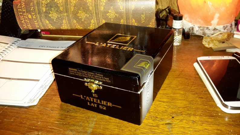May include: A black wooden box with gold accents and the text "L'Atelier 15 Lat 52" on the front. The box is closed and has a glossy finish.