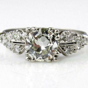 Könnte beinhalten: Ein Verlobungsring im Vintage-Stil mit einem großen, rund geschliffenen Diamanten als Mittelpunkt. Kleinere Diamanten flankieren den zentralen Stein, der in einem silbernen oder weißgoldenen Band gefasst ist. Der Ring hat aufwendige Details.