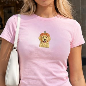 Pode incluir: Uma t-shirt de manga curta em rosa claro com um bordado central de um cão golden retriever a usar uma abóbora na cabeça. A peça é combinada com uma mala de ombro branca e calças de ganga azuis com um botão prateado visível.
