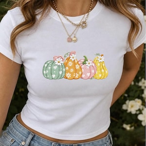 Pode incluir: Uma t-shirt branca de manga curta com uma estampa gráfica de quatro abóboras em tons de verde, laranja, rosa e amarelo. Cada abóbora é decorada com pequenos padrões de corações e margaridas brancas, encimada por uma única margarida.