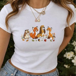 Könnte beinhalten: Ein weißes T-Shirt mit kurzen Ärmeln und einem farbenfrohen Grafikdruck, der eine Eule, zwei Füchse und einen Jagdhund zeigt, die beieinander sitzen. Die Illustration enthält kleine Herbstblätter, Blumen und einen Pilz.