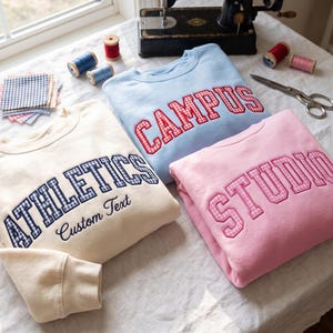 Può includere: Tre felpe girocollo color crema, azzurro e rosa. La felpa color crema presenta la scritta ATHLETICS in applicazione a quadretti blu con il testo Custom Text sottostante. La felpa azzurra riporta CAMPUS in quadretti rossi, quella rosa STUDIO in quadretti rosa.