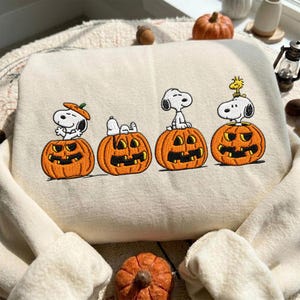 May include: A cream-colored sweatshirt featuring an embroidered design of four orange jack-o'-lanterns. Each pumpkin contains a white Snoopy character in different poses, with one pumpkin topped by a small yellow Woodstock bird figure.