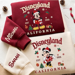 Può includere: Una felpa bordeaux con ricamato Topolino con cappello di Natale e la scritta Disneyland California Est. 1955 con il nome Michael sulla manica, e una felpa color crema con Minnie e il nome Sarah ricamato sulla manica.
