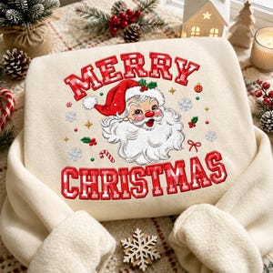 Könnte beinhalten: Ein cremefarbenes Sweatshirt mit einer zentralen Stickerei eines weihnachtlichen Santa-Claus-Gesichts im Vintage-Stil mit roter Mütze. Der Schriftzug MERRY CHRISTMAS ist in rot-weiß karierten Buchstaben über und unter dem Motiv gestickt.