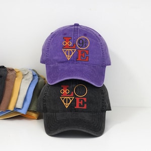 Op de afbeelding: Twee baseballpetten, één paars en één zwart, op elkaar gestapeld. Elke pet heeft een geborduurd "LOVE"-ontwerp met een afbeelding. De petten zijn gemaakt van een zachte, versleten stof. Andere petten zijn zichtbaar op de achtergrond.