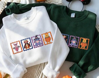 Puede incluir: Una sudadera blanca y una sudadera verde oscuro, ambas con una fila horizontal de cuatro marcos rectangulares que contienen ilustraciones de Piglet como murciélago, Tigger como vampiro, Igor como bruja y Winnie the Pooh como calabaza.