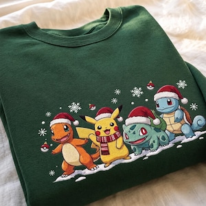 Pode incluir: Uma camisola de gola redonda verde-escura com uma impressão gráfica de quatro personagens a usar gorros de Pai Natal vermelhos. As figuras incluem um lagarto laranja, um rato amarelo com um cachecol vermelho, uma criatura verde e uma tartaruga.