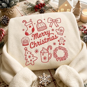 Könnte beinhalten: Ein cremefarbenes Sweatshirt mit roten, gestickten Weihnachtsmotiven wie einem Weihnachtsstrumpf, einer Mütze, einer Schleife, einem Fäustling, einer Schneekugel, einem Lebkuchenmann, einem Tannenbaum, einer Tasse mit Zuckerstange und einem Kranz.