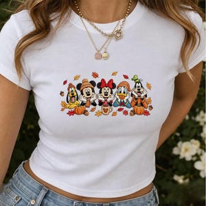 Może przedstawiać: Biały t-shirt z krótkim rękawem z nadrukiem graficznym przedstawiającym Pluto, Myszkę Miki, Myszkę Minnie, Kaczora Donalda i Goofy'ego w jesiennych czapkach i szalikach, w otoczeniu pomarańczowych dyń, brązowych żołędzi i kolorowych liści.
