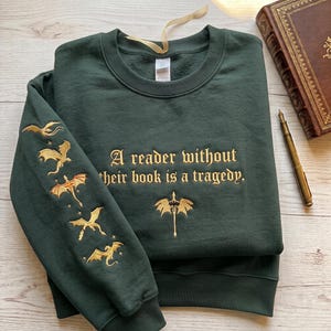 May include: A dark green crewneck sweatshirt featuring gold embroidery. The chest displays the text "A reader without their book is a tragedy." with a sword icon below. The left sleeve features four embroidered gold dragons in flight.