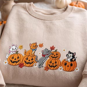 Könnte beinhalten: Ein beigefarbenes Sweatshirt mit Rundhalsausschnitt und einer Stickerei von vier Kätzchen, die mit orangefarbenen Kürbissen spielen. Die Kätzchen sind weiß, orange, grau und schwarz und mit kleinen Herbstblättern sowie weißen Gänseblümchen verziert.