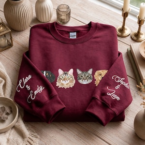 Op de afbeelding: Een bordeauxrode sweater met ronde hals en een rij van vier geborduurde dierenportretten op de borst. Op de linkermouw staan de namen Clara & Emily geborduurd en op de rechtermouw de namen Sophia & Liam in het wit.