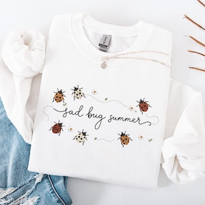 Può includere: Felpa bianca con la scritta "sad bug summer" in corsivo nero, decorata con illustrazioni di coccinelle e piccoli fiori bianchi. Una collana d'argento con un piccolo pendente è drappeggiata sulla felpa.