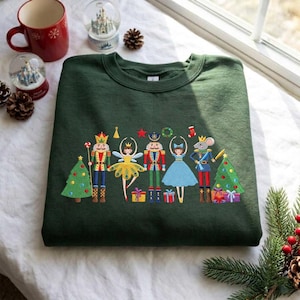 Può includere: Una felpa girocollo verde scuro con una grafica ricamata a tema Schiaccianoci. Il design include uno schiaccianoci, una fata, una ballerina e un topo circondati da due alberi di Natale decorati e scatole regalo colorate.
