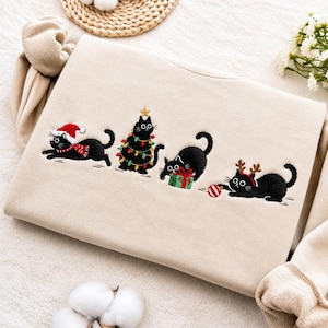 Può includere: Una felpa beige con un ricamo raffigurante quattro gatti neri. Un gatto indossa un cappello da Babbo Natale e una sciarpa, uno è avvolto da luci natalizie colorate, uno gioca con un pacchetto regalo verde e uno indossa corna da renna.