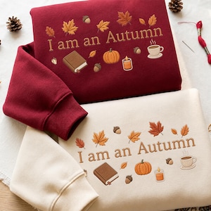 Op de afbeelding: Een donkerrood sweatshirt en een crèmekleurig sweatshirt, elk voorzien van geborduurde herfstmotieven zoals esdoornbladeren, eikels, een boek, een pompoen, een kaars en een koffiekopje, met de tekst "I am an Autumn" geborduurd in bruin garen.