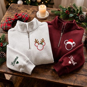 Könnte beinhalten: Ein hellgraues Sweatshirt mit Viertelreißverschluss, bestickt mit einem Maus-Symbol mit Rentiergeweih und dem Namen Emma am Ärmel, sowie ein kastanienbraunes Sweatshirt mit einem Maus-Symbol mit Weihnachtsmütze und dem Namen Noah am Ärmel.