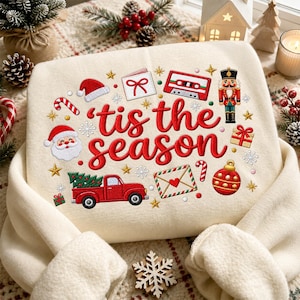 Könnte beinhalten: Ein cremefarbenes Sweatshirt mit roter Stickerei, die den Schriftzug 'tis the season' zeigt, umgeben von festlichen Symbolen wie einem roten Lastwagen mit Tannenbaum, einem Nussknacker, einer Kassette, einem Weihnachtsmann und einer roten Kugel.