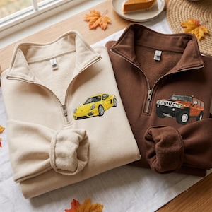 Könnte beinhalten: Zwei Sweatshirts mit kurzem Reißverschluss, eines in Creme und eines in Braun. Das cremefarbene Sweatshirt zeigt einen gestickten gelben Sportwagen, während das braune Sweatshirt einen gestickten orangefarbenen SUV aufweist. Beide haben lange Ärmel.