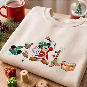 Könnte beinhalten: Ein cremefarbenes Sweatshirt mit Rundhalsausschnitt und einer Stickerei von Mickey Mouse als Zauberlehrling in einem roten Gewand mit weißem Besatz und grünem Hut, dazu ein Zauberstab, ein Besen, zwei Eimer und ein offenes Buch mit Glocken.