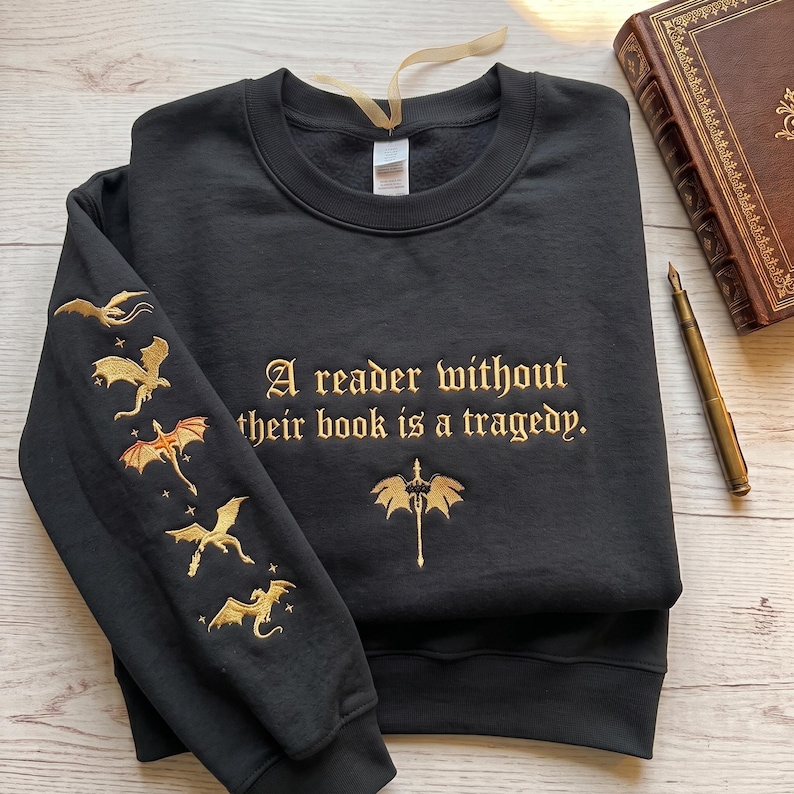 May include: A black crewneck sweatshirt featuring gold embroidery. The chest displays the text "A reader without their book is a tragedy." with a small dragon icon below. The left sleeve features a vertical row of four embroidered gold dragons.