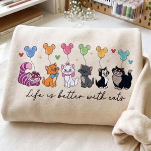 Könnte beinhalten: Ein cremefarbenes Sweatshirt mit einem Grafikdruck von sechs Zeichentrickkatzen, die jeweils einen bunten Luftballon in Form von Mausohren halten. Der Text "Life is better with cats" ist in schwarzer Schreibschrift unter den Katzen gedruckt.