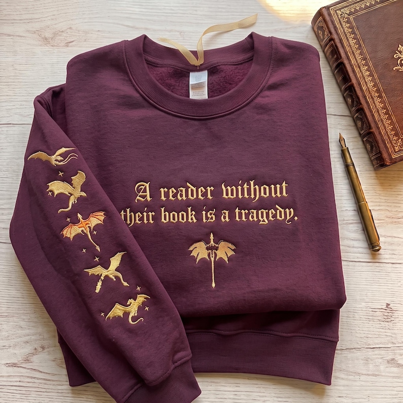 May include: A deep burgundy crewneck sweatshirt featuring gold embroidery. The chest displays the text A reader without their book is a tragedy. with a dragon emblem below. The left sleeve features five embroidered gold dragons in flight.