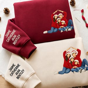 Può includere: Una felpa bordeaux e una felpa color crema, entrambe con la grafica di una famiglia di quattro persone che indossano maglioni rossi e cappelli di Babbo Natale, formando un tetto con le braccia. Le maniche riportano i nomi Jaime, Catherine, Avni, Bentley.
