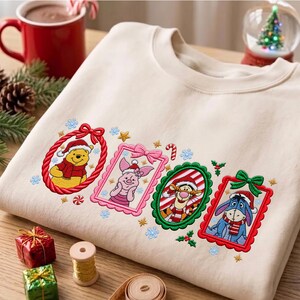 Può includere: Una felpa girocollo beige con ritratti ricamati di Winnie the Pooh, Pimpi, Tigro e Ih-Oh, ciascuno incorniciato in colori festivi con fiocchi. I personaggi indossano cappelli da Babbo Natale e sciarpe, circondati da piccoli fiocchi di neve e stelle.