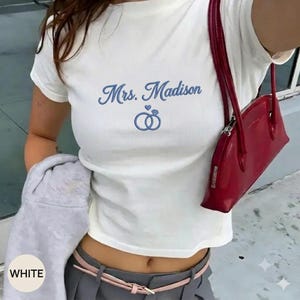 Può includere: Una t-shirt bianca a maniche corte in tessuto a coste con il testo ricamato in blu Mrs. Madison sopra un grafico di due anelli intrecciati e un piccolo cuore. Una borsa a spalla rossa con chiusura a zip è indossata sulla spalla.