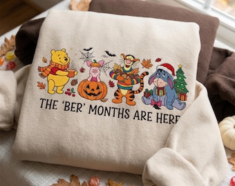 Puede incluir: Una sudadera beige con un gráfico de Winnie the Pooh, Piglet en una calabaza, Tigger y Eeyore con un árbol de Navidad. El diseño incluye hojas de otoño, una telaraña y el texto THE 'BER' MONTHS ARE HERE en letras de color negro.