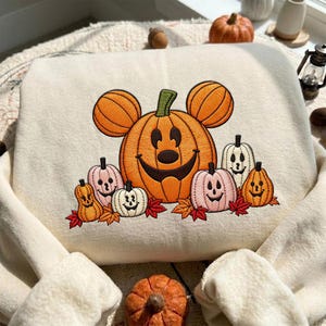 Peut inclure: Un sweat-shirt couleur crème avec une broderie représentant une grande citrouille orange en forme de tête de souris souriante, entourée de citrouilles plus petites dans des tons orange, rose et blanc, agrémentées de feuilles rouges et orange.