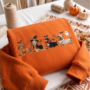 Könnte beinhalten: Ein orangefarbenes Sweatshirt mit einer Stickerei von vier Dackeln in Halloween-Kostümen. Die Hunde sind als Kürbis, Hexe, Vampir und Mumie verkleidet, umgeben von kleinen gestickten Fledermäusen, Spinnennetzen und einem Geist.