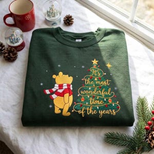 Könnte beinhalten: Ein dunkelgrünes Sweatshirt mit Rundhalsausschnitt, das einen gestickten gelben Bären mit rot-weiß gestreiftem Schal neben einem grünen Weihnachtsbaum zeigt, mit dem goldenen Text, It's the most wonderful time of the years.
