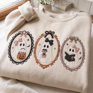 Könnte beinhalten: Ein beigefarbenes Sweatshirt mit Rundhalsausschnitt und drei gestickten ovalen Rahmen. Jeder Rahmen zeigt ein weißes Gespenst mit einer Schleife. Das erste Gespenst hält einen Kürbis, das zweite einen Kaffeebecher und das dritte ein offenes Buch.