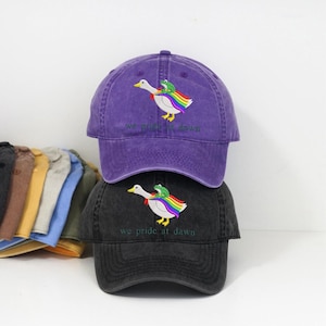 Può includere: Due cappellini da baseball, uno viola e uno nero, impilati l'uno sull'altro. Ogni cappellino presenta un disegno ricamato di un'anatra bianca con un'ala arcobaleno e una rana verde. Il testo "we pride at dawn" è ricamato sotto.
