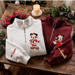 Op de afbeelding: Een lichtgrijs sweatshirt met kwartrits, voorzien van een geborduurde Mickey Mouse in kerstmanpak en de naam Noah op de mouw, naast een bordeauxrood sweatshirt met een geborduurde Minnie Mouse en de naam Sophia op de mouw.