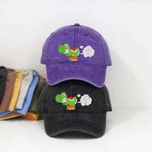 May include: Two baseball caps, one purple and one black, stacked on top of each other. Each cap features an embroidered green dinosaur character with a red saddle and white cloud-like accents. The caps have a curved brim.